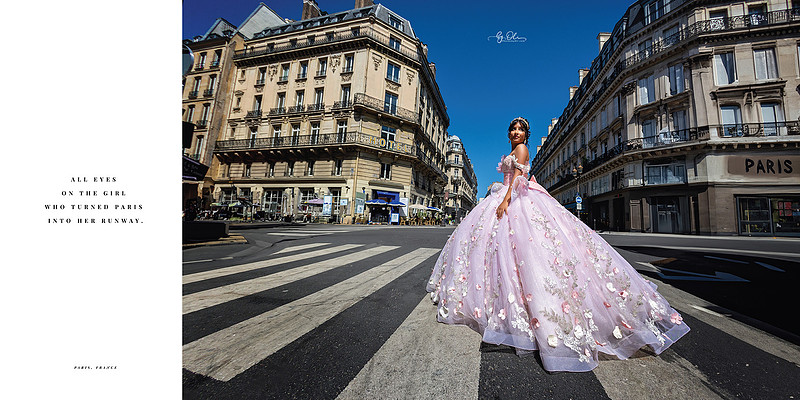 Quinceañera girl in a pink floral gown standing in the middle of a Parisian street with classic Haussmann buildings in the background, posing as if on a fashion runway.