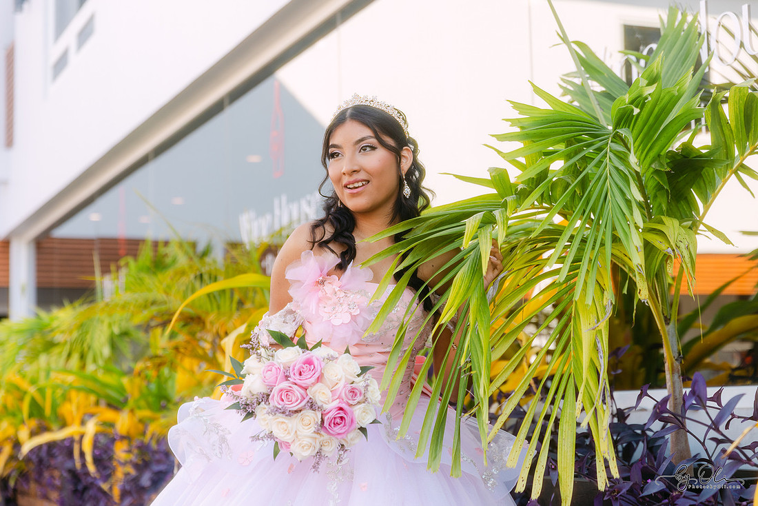 Chelsea Monique Alfaro outdoors during her quinceañera in Belize, holding a bouquet and standing among palm trees in warm tropical light.