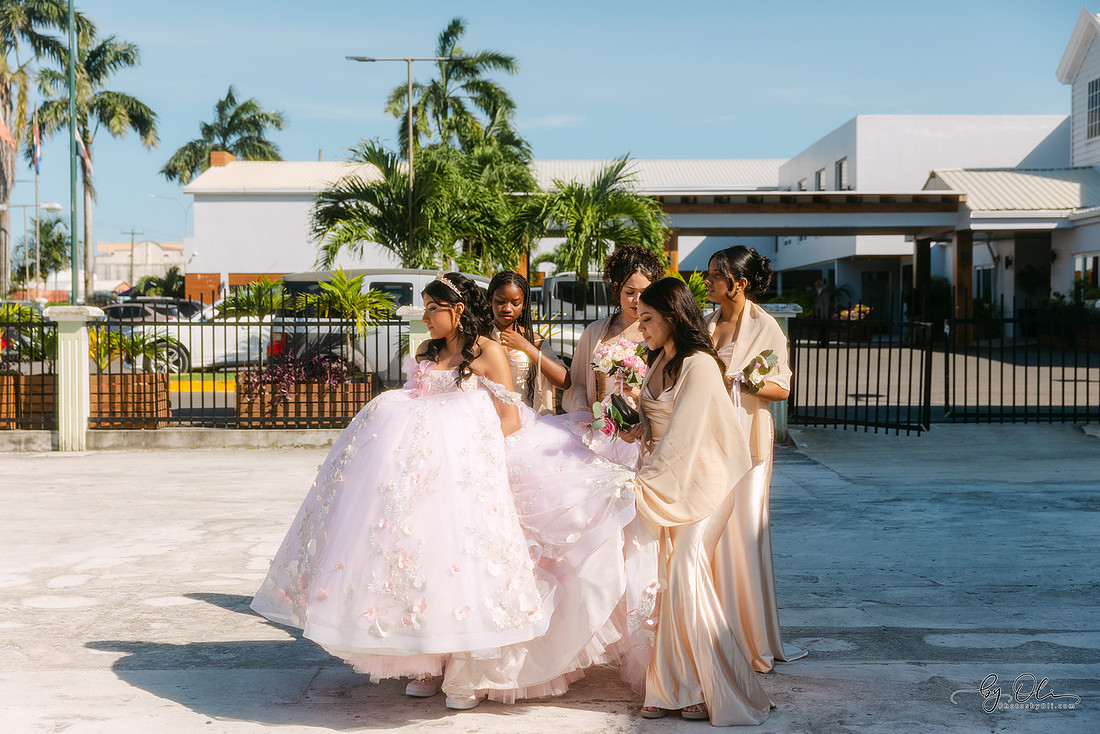 Chelsea Monique Alfaro surrounded by her friends as they help her walk toward the party venue before the quinceañera celebration in Belize, adjusting her dress and sharing a supportive moment.