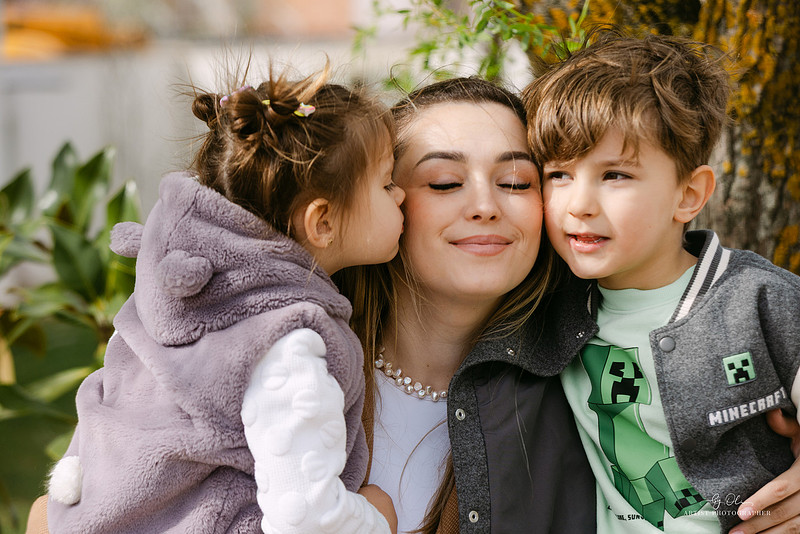 
Mama cu doi copii mici langa ea, fetita sarutand-o pe obraz si baiat privind direct in camera, portret emotional de familie, Photos by Oli Timisoara